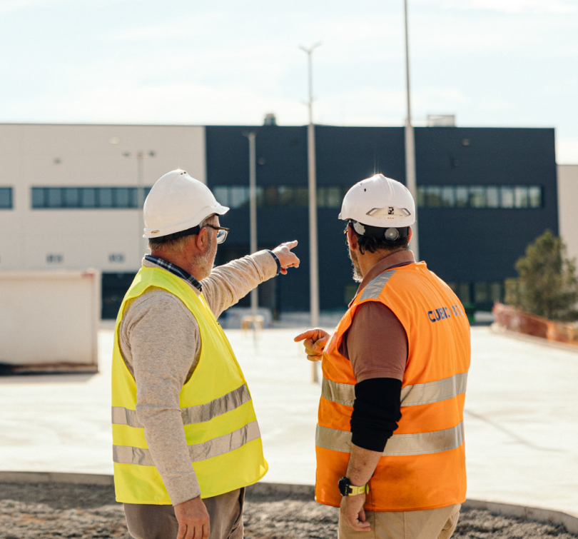 Dos trabajadores con chaleco y casco supervisan la construcción de una nave industrial.